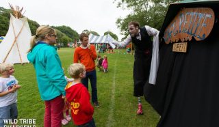 A performer in costume entertains a family with two children outside a tent labelled Café Petite at an outdoor festival. Other people and tents are visible in the grassy background. Text reads: Wild Tribe Children’s Festival.