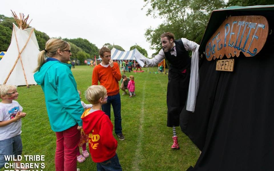 A performer in costume entertains a family with two children outside a tent labelled Café Petite at an outdoor festival. Other people and tents are visible in the grassy background. Text reads: Wild Tribe Children’s Festival.