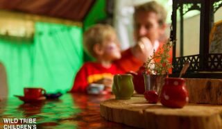 A blurred adult and child sit at a table set with colourful toy tea cups and a teapot. The image has a warm, playful atmosphere. Wild Tribe Childrens Festival is written in white text in the bottom left corner.