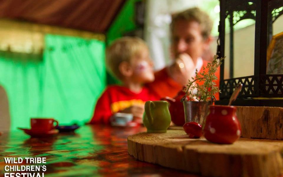 A blurred adult and child sit at a table set with colourful toy tea cups and a teapot. The image has a warm, playful atmosphere. Wild Tribe Childrens Festival is written in white text in the bottom left corner.