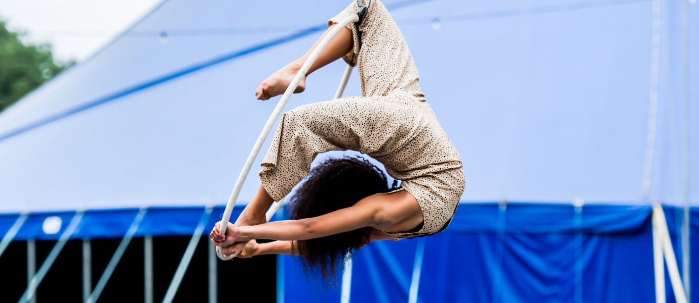 Aerial performer in a beige outfit hanging upside down from a white rope, with legs hooked and arms extended, in front of a large blue circus marquee.