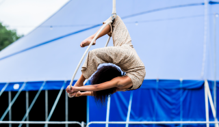 Aerial performer in a beige outfit hanging upside down from a white rope, with legs hooked and arms extended, in front of a large blue circus marquee.