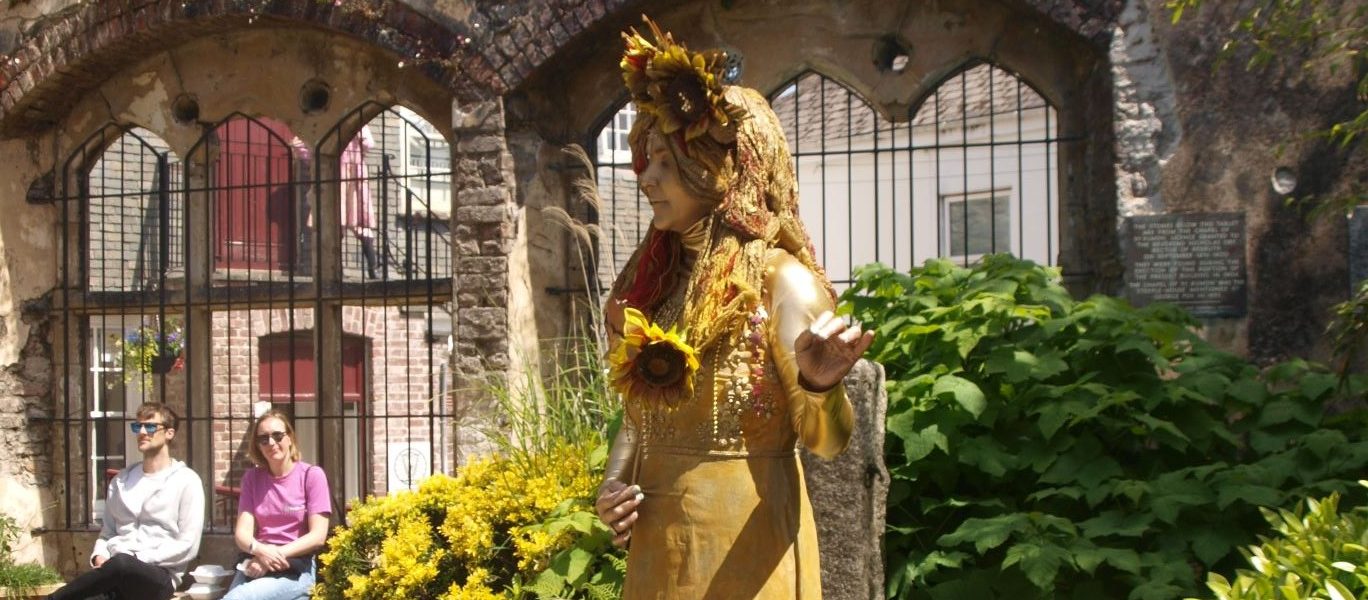A golden statue of a woman holding a sunflower stands in a garden with lush green bushes and yellow flowers, in front of ancient stone arches. Two people sit on a bench in the background, enjoying the sunny day.