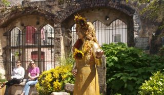 A golden statue of a woman holding a sunflower stands in a garden with lush green bushes and yellow flowers, in front of ancient stone arches. Two people sit on a bench in the background, enjoying the sunny day.