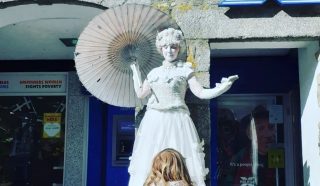 A street performer dressed as a white statue with a parasol stands on a plinth, posing outside a shop. A child with red hair stands in front of the performer, looking up in wonder.