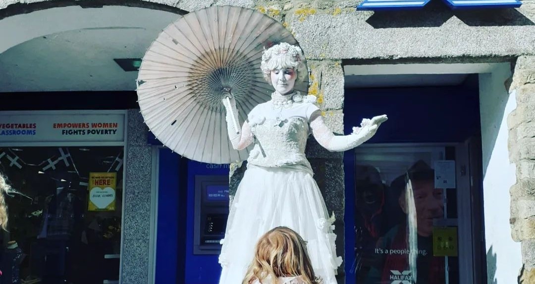 A street performer dressed as a white statue with a parasol stands on a plinth, posing outside a shop. A child with red hair stands in front of the performer, looking up in wonder.
