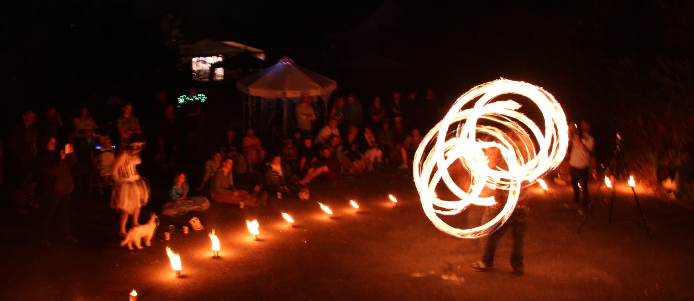 A fire performer spins flaming poi, creating swirling light patterns at night. A crowd sits and stands in a semicircle, watching the show. Small flames line the ground, and a dog is present among the spectators.