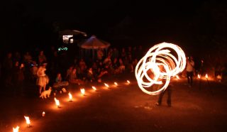 A fire performer spins flaming poi, creating swirling light patterns at night. A crowd sits and stands in a semicircle, watching the show. Small flames line the ground, and a dog is present among the spectators.
