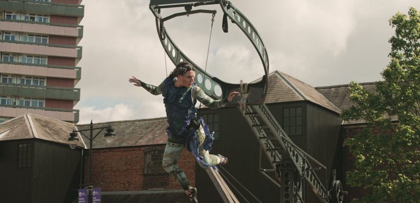 A performer in a blue costume balances on a curved metal structure outdoors, with brick buildings and a tall block of flats in the background. Trees and cloudy sky are also visible.