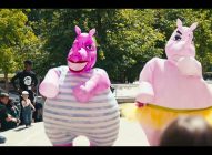 Two people in colourful, oversized hippo costumes—one pink with stripes, one pink with a yellow tutu—dance outdoors while a crowd of people watches and takes photos. Trees and sunlight are visible in the background.