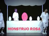 Two performers in white outfits and pink glasses stand on a stage with a white booth labelled MONSTRUO ROSA. Behind the booth are four white egg shapes and one large pink egg at the centre.