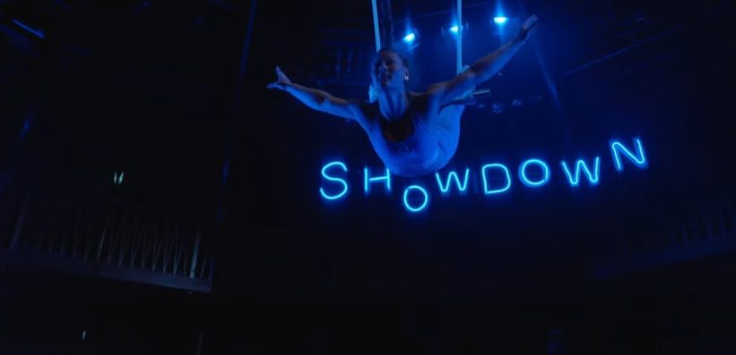 An aerial performer suspended mid-air under blue stage lights, arms outstretched, with the word SHOWDOWN illuminated in neon letters behind them.