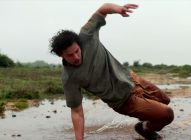 A man with curly hair slips and falls in a muddy puddle outdoors, his arms flailing as he loses balance. The sky is overcast and the ground is wet, with green bushes in the background.