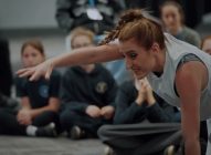 A woman in costume performs an expressive, dramatic gesture with one arm extended, while seated children in school uniforms watch her intently in the background.