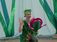 A person dressed in a green leafy costume waves and holds a large pink and red flower prop, performing in front of a backdrop decorated with tall, green plant shapes.