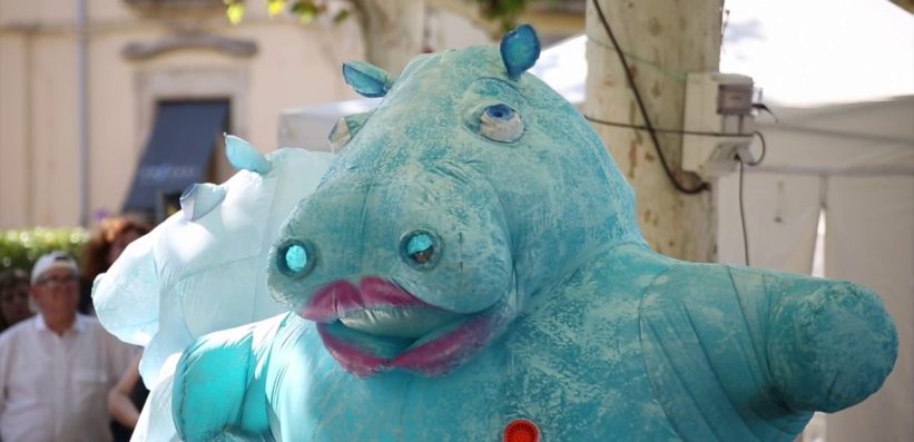A person in a large, blue hippopotamus costume with pink lips and glowing blue eyes stands outdoors at an event, with trees and people in the background.