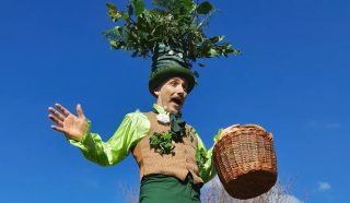 A performer in a green and brown costume with leaves and a large plant hat gestures with one hand and holds a wicker basket in the other, standing against a clear blue sky.