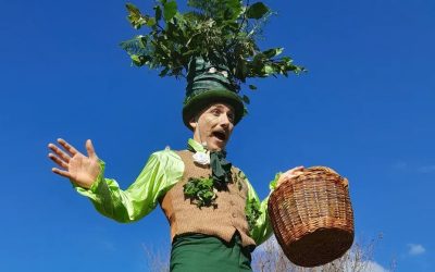 A performer in a green and brown costume with leaves and a large plant hat gestures with one hand and holds a wicker basket in the other, standing against a clear blue sky.