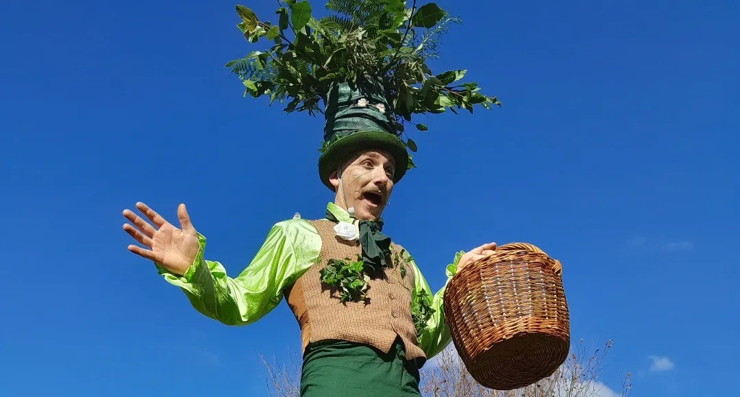 A performer in a green and brown costume with leaves and a large plant hat gestures with one hand and holds a wicker basket in the other, standing against a clear blue sky.