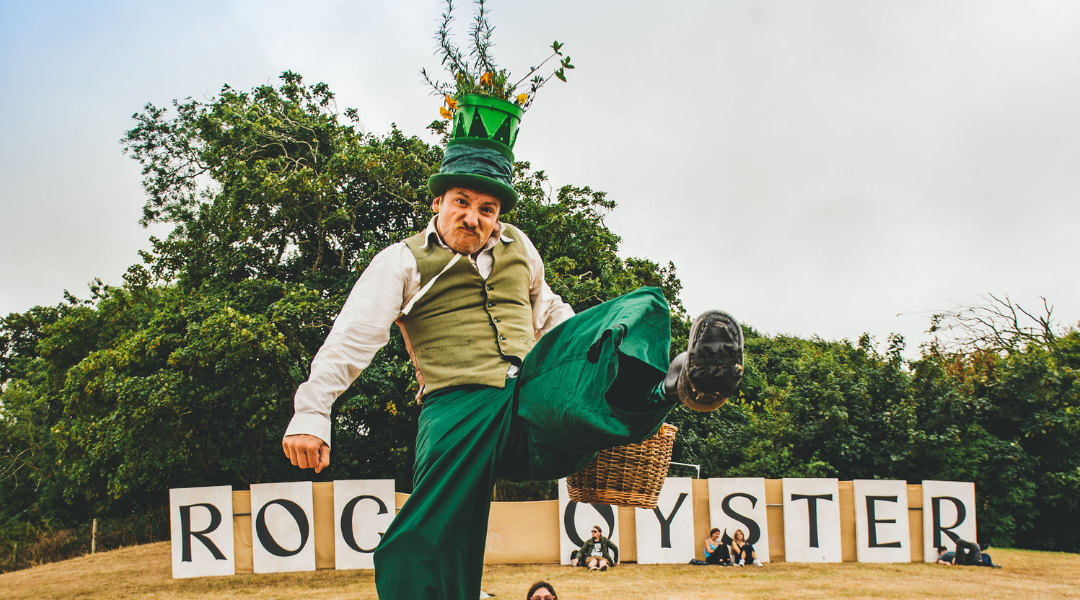 A performer in green trousers and a green waistcoat kicks one leg up, wearing a tall hat decorated with a plant. Large letters behind him spell ROCK OYSTER at an outdoor event with people sitting on grass.