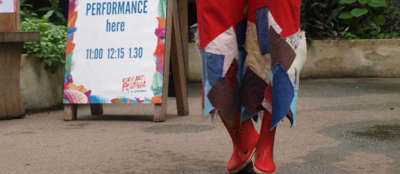 Person in red boots and colourful patchwork trousers walking towards a sign that reads Hotch Potch Performance here 11:00 12:15 13:30 at an outdoor event with greenery in the background.