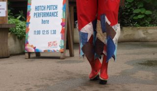 Person in red boots and colourful patchwork trousers walking towards a sign that reads Hotch Potch Performance here 11:00 12:15 13:30 at an outdoor event with greenery in the background.