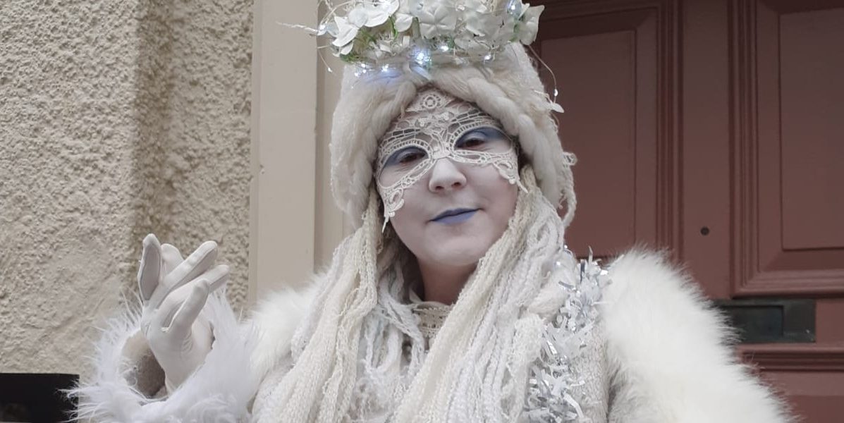 A person dressed in an elaborate winter-themed costume with white make-up, a lace mask, white fur coat, knitted accessories, and a crown made of flowers and lights, standing in front of a red door.
