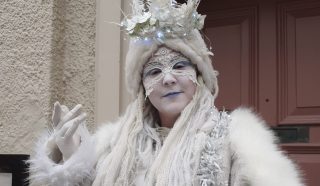 A person dressed in an elaborate winter-themed costume with white make-up, a lace mask, white fur coat, knitted accessories, and a crown made of flowers and lights, standing in front of a red door.