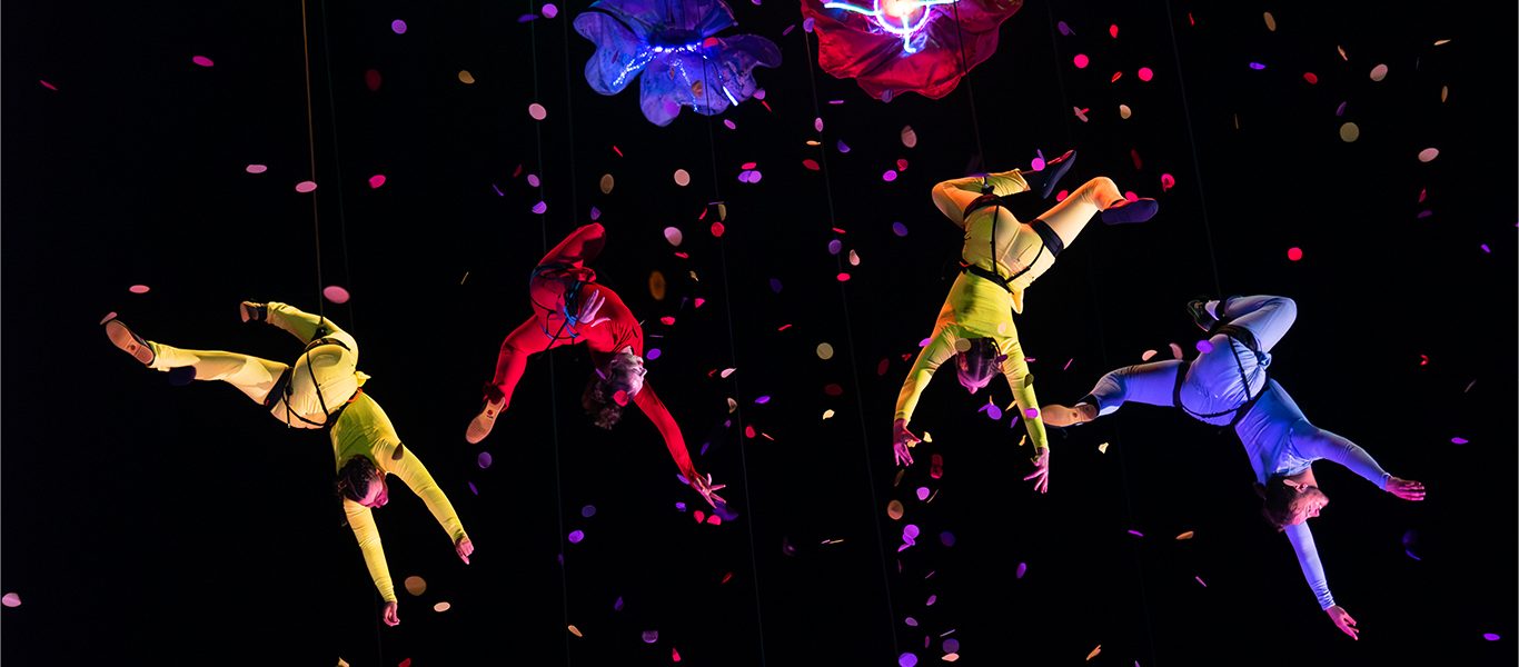 Acrobats in colourful suits hang suspended in mid-air, performing under bright lights with flower-shaped props above them, surrounded by falling confetti against a dark background.