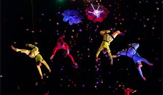 Acrobats in colourful suits hang suspended in mid-air, performing under bright lights with flower-shaped props above them, surrounded by falling confetti against a dark background.