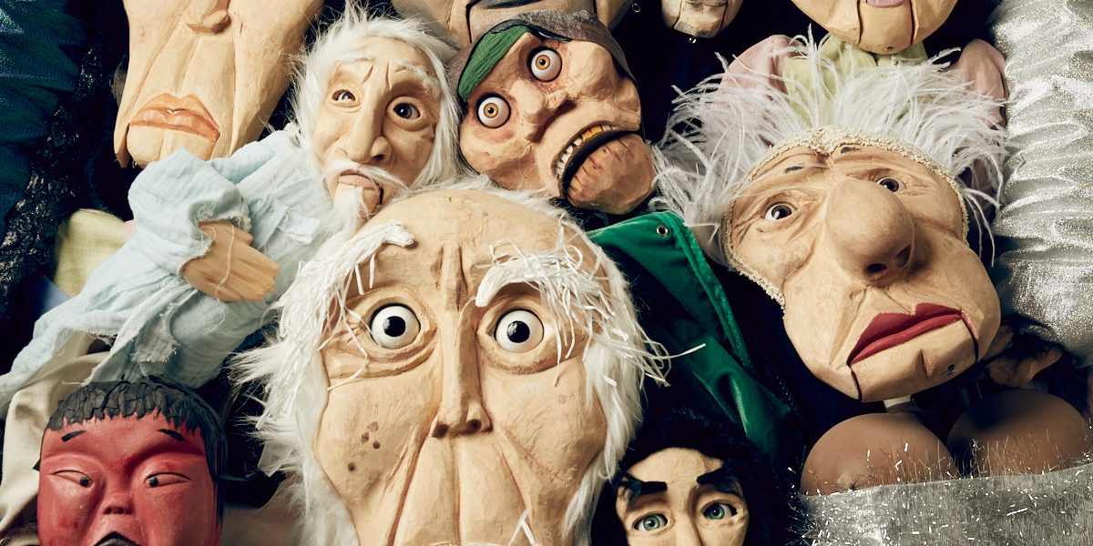 A group of colourful puppets with expressive faces clustered together, featuring various characters including an elderly man, a devil, and others with distinct costumes and exaggerated features.