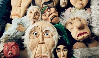 A group of colourful puppets with expressive faces clustered together, featuring various characters including an elderly man, a devil, and others with distinct costumes and exaggerated features.