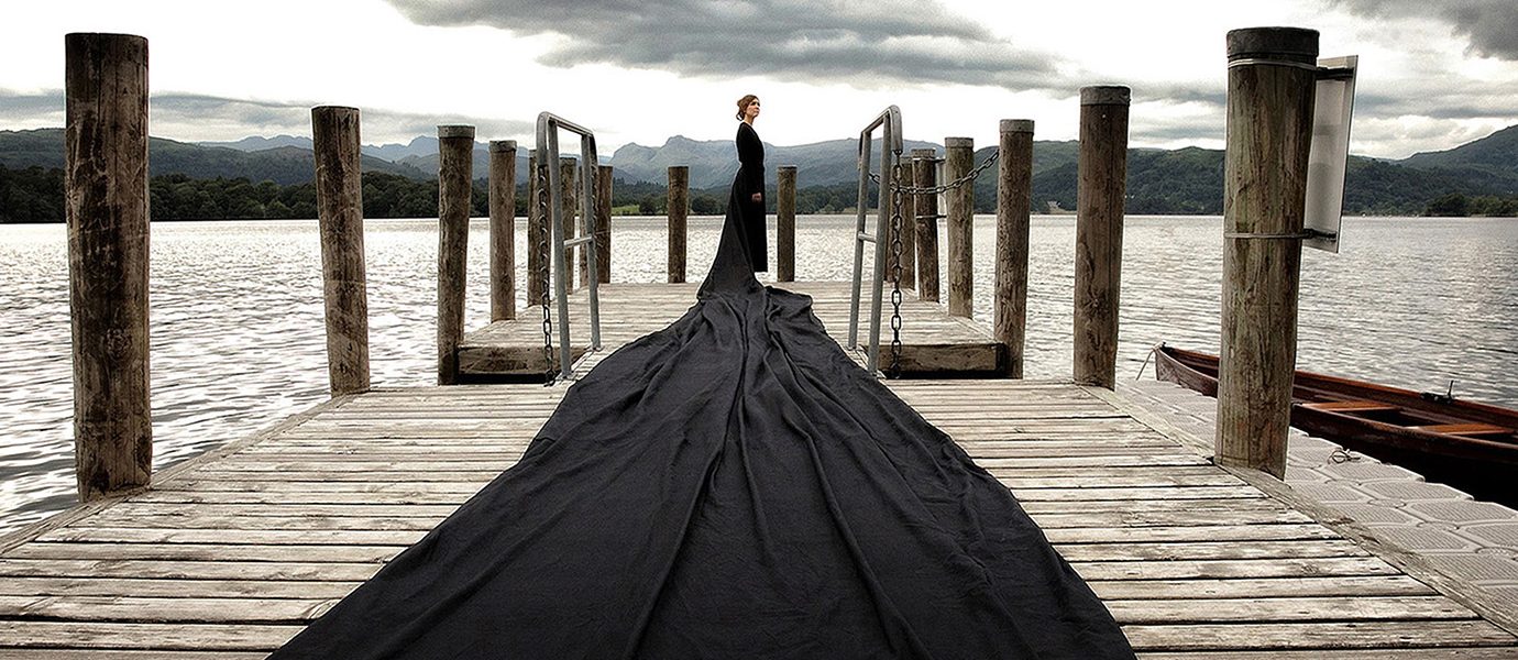 A person in black stands on a wooden jetty by a lake, wearing a dress with an extremely long train that flows across the jetty. The sky is cloudy and mountains are visible in the background.