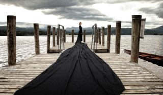 A person in black stands on a wooden jetty by a lake, wearing a dress with an extremely long train that flows across the jetty. The sky is cloudy and mountains are visible in the background.