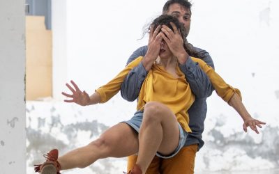 A man stands behind a woman, covering her eyes with his hands. She extends one leg forward and her arms out, wearing a yellow top and denim skirt. The background is a white, textured wall.