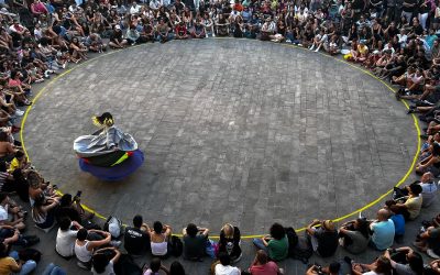 A large crowd sits in a circle on grey pavement, watching a single performer in colourful attire spinning in the centre. The scene is lively and outdoors, with spectators closely watching the performance.