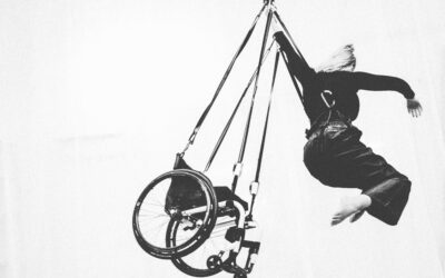 A person with a wheelchair is suspended in mid-air by harnesses, reaching upward with one arm. The background is plain and white, creating a sense of openness.