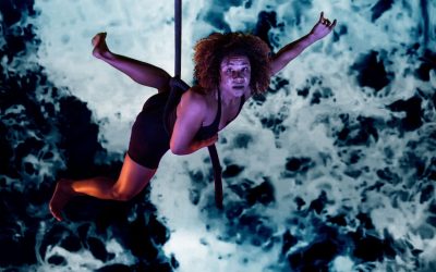 A person with curly hair performs an aerial act on a rope, wearing a black outfit, against a blue and black abstract background.
