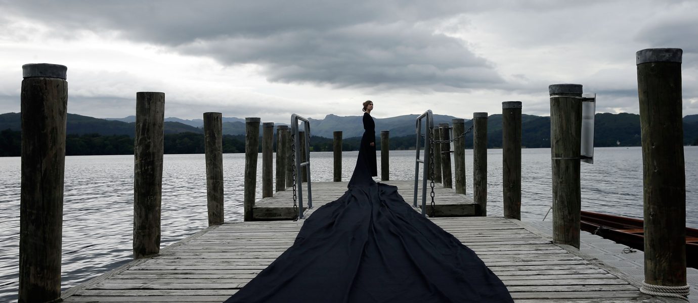 A person in a long, flowing black dress stands at the end of a wooden jetty over a lake, with mountains and cloudy skies in the background.
