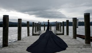 A person in a long, flowing black dress stands at the end of a wooden jetty over a lake, with mountains and cloudy skies in the background.