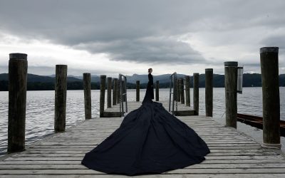 A person in a long, flowing black dress stands at the end of a wooden jetty over a lake, with mountains and cloudy skies in the background.