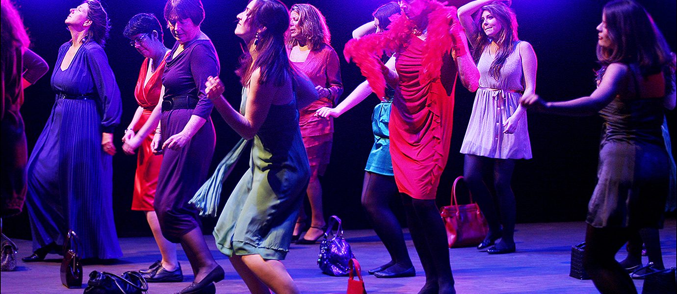 A group of women in colourful dresses dance enthusiastically on a stage with purple lighting, handbags on the ground, and bright lights shining behind them.