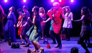 A group of women in colourful dresses dance enthusiastically on a stage with purple lighting, handbags on the ground, and bright lights shining behind them.