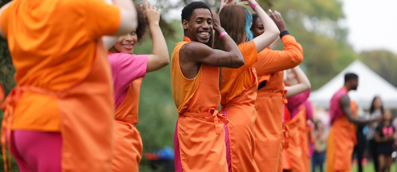 A group of people wearing matching orange and pink outfits dance outdoors in a line, smiling and raising their arms. The background is grassy with trees and a white marquee visible.