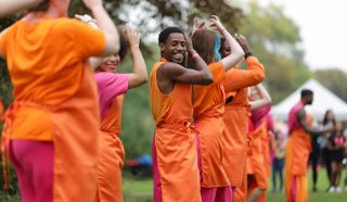 A group of people wearing matching orange and pink outfits dance outdoors in a line, smiling and raising their arms. The background is grassy with trees and a white marquee visible.