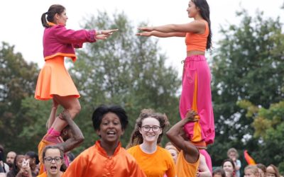 A group of performers in vibrant orange and pink outfits participate in an outdoor event. Two people are balanced atop others’ shoulders, whilst others smile and move forwards. A crowd watches in the background.