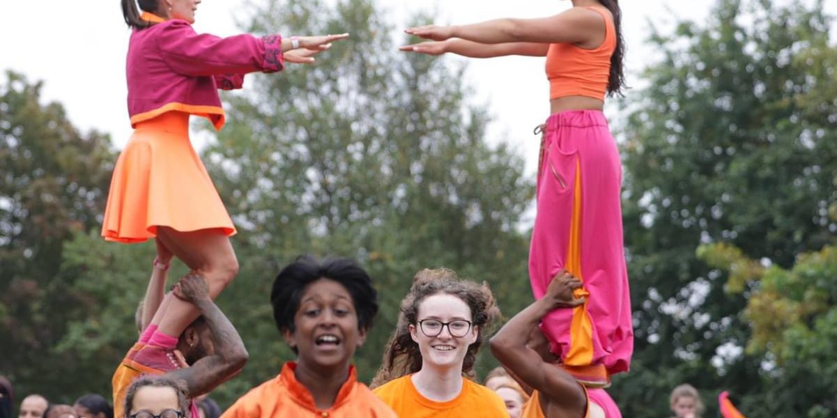 A group of performers in bright orange and pink outfits form human pyramids outdoors, with two girls balanced on others’ shoulders. A crowd watches in the background. The mood is lively and energetic.
