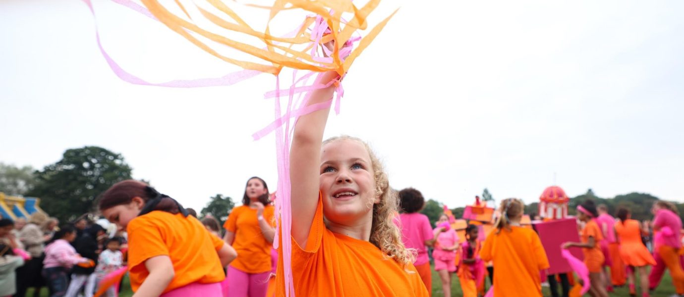 A young girl in an orange shirt smiles and waves orange and pink streamers in the air, surrounded by a crowd of people dressed in orange and pink outfits at an outdoor event.