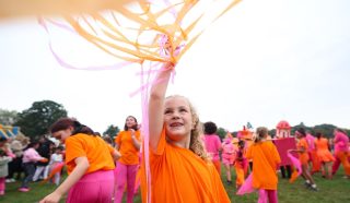 A young girl in an orange shirt smiles and waves pink and yellow streamers above her head. She is outdoors with a crowd of people, many also wearing orange and pink, on a grassy field during a festive event.