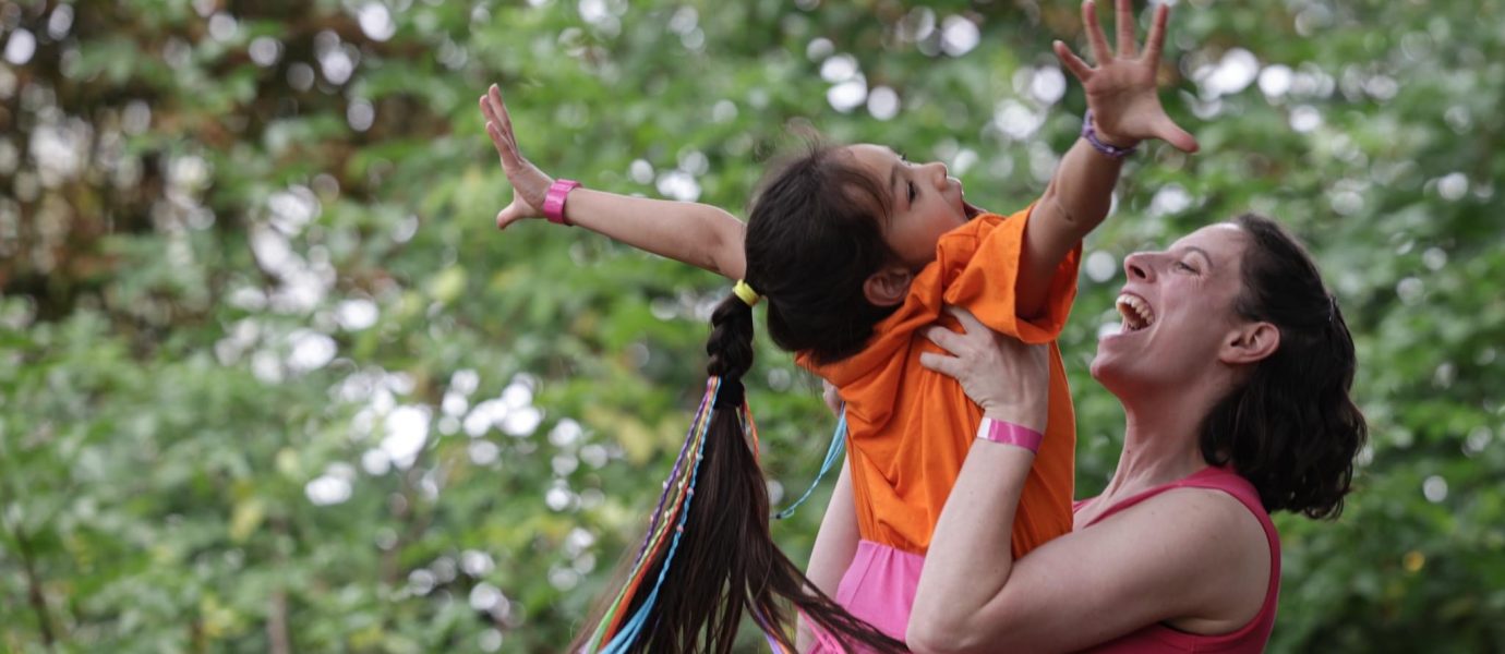 A woman joyfully lifts a smiling girl with a long ponytail and colourful ribbons into the air outdoors, both reaching their arms upward against a background of green trees.